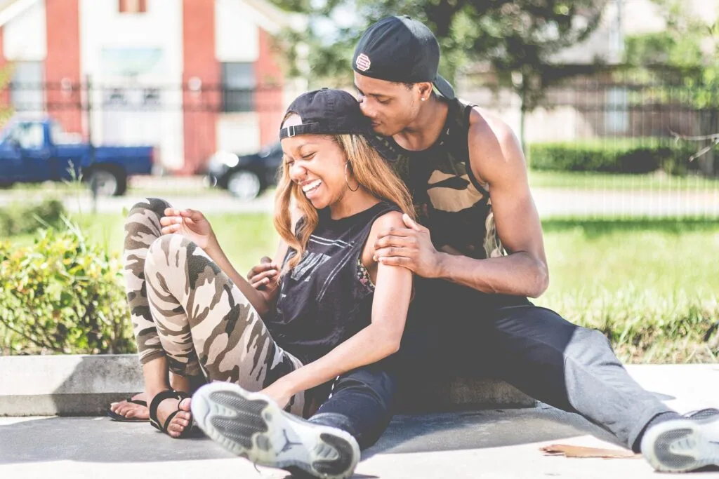 A man and woman in baseball caps laugh together while sitting outdoors on pavement, with a red and white building in the background.