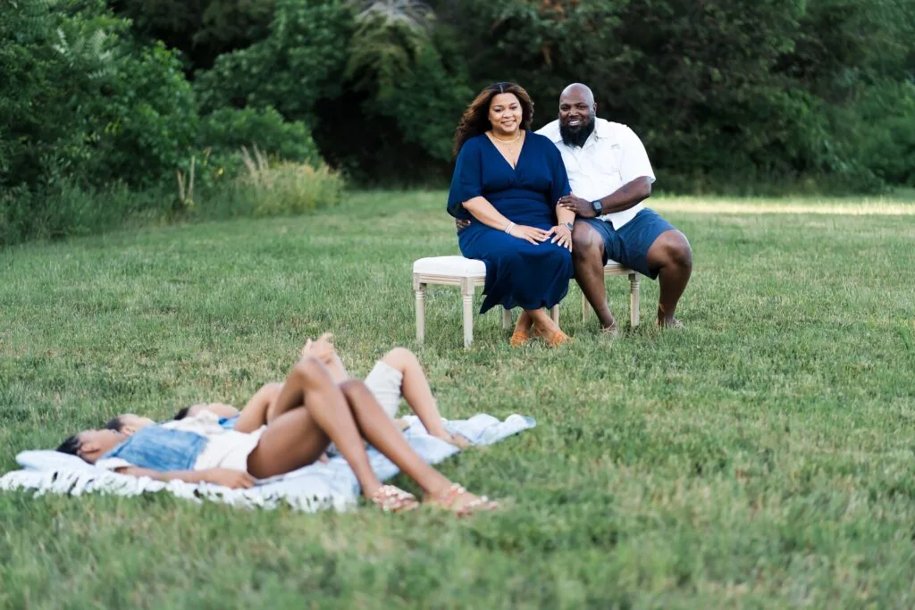Two adults sitting on stools watching a child lying on a blanket in a grassy outdoor area with trees in background