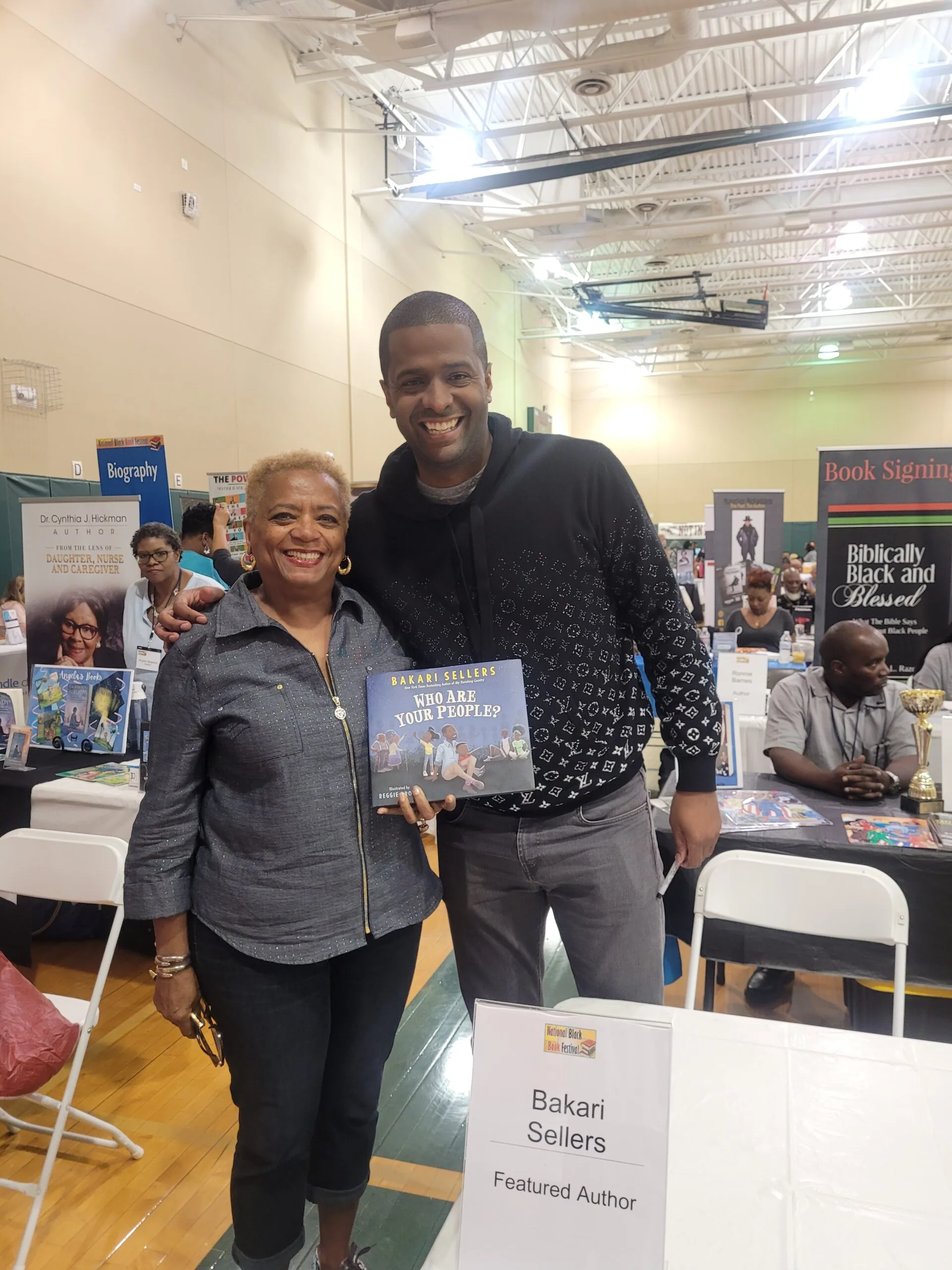 Two people smiling at a book signing event, with one holding a book titled 'You Are Your Child's First Teacher', at an indoor venue with tables and bookshelves visible in background