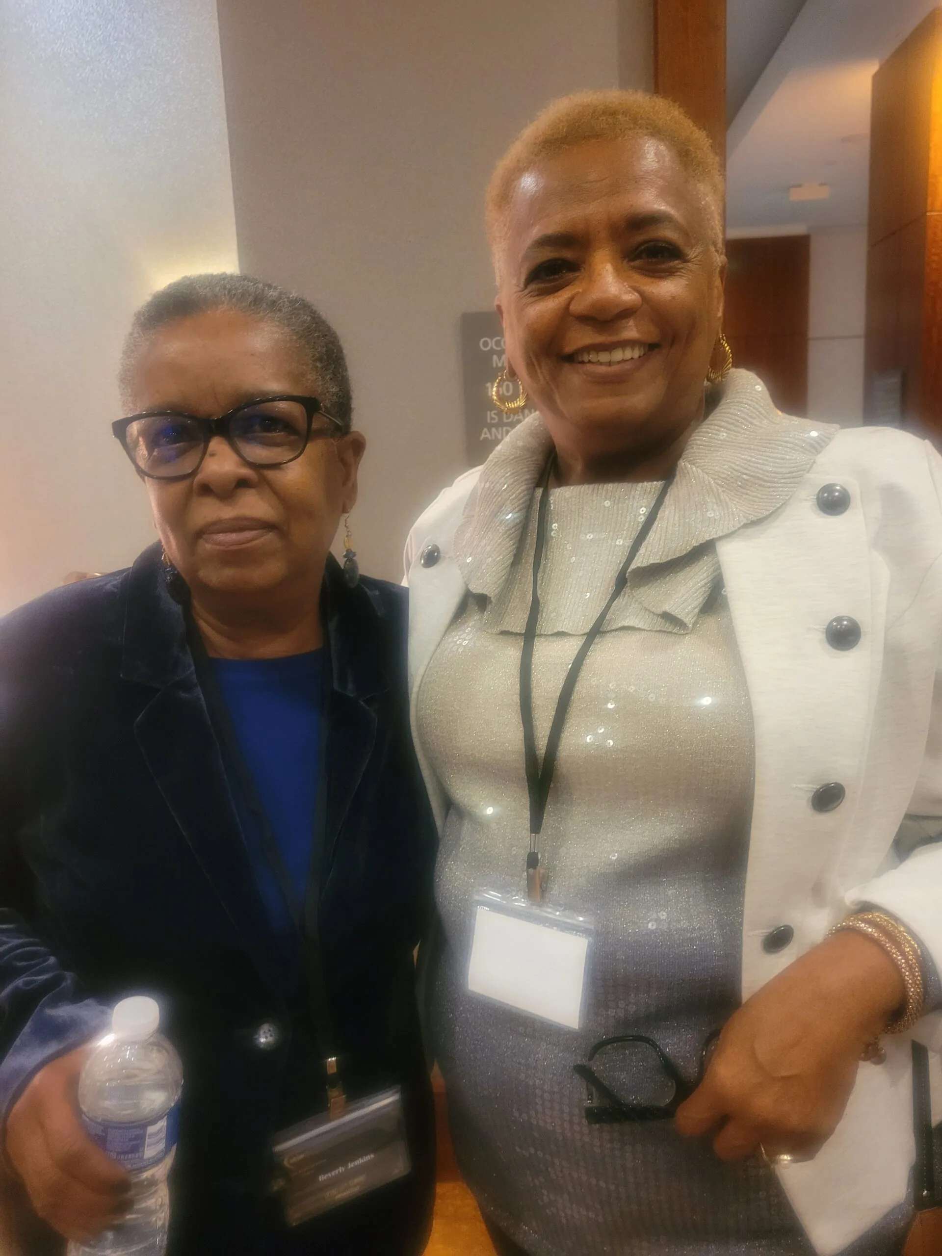 Two women posing together indoors, one wearing glasses and dark jacket, the other wearing white blazer with credential badge