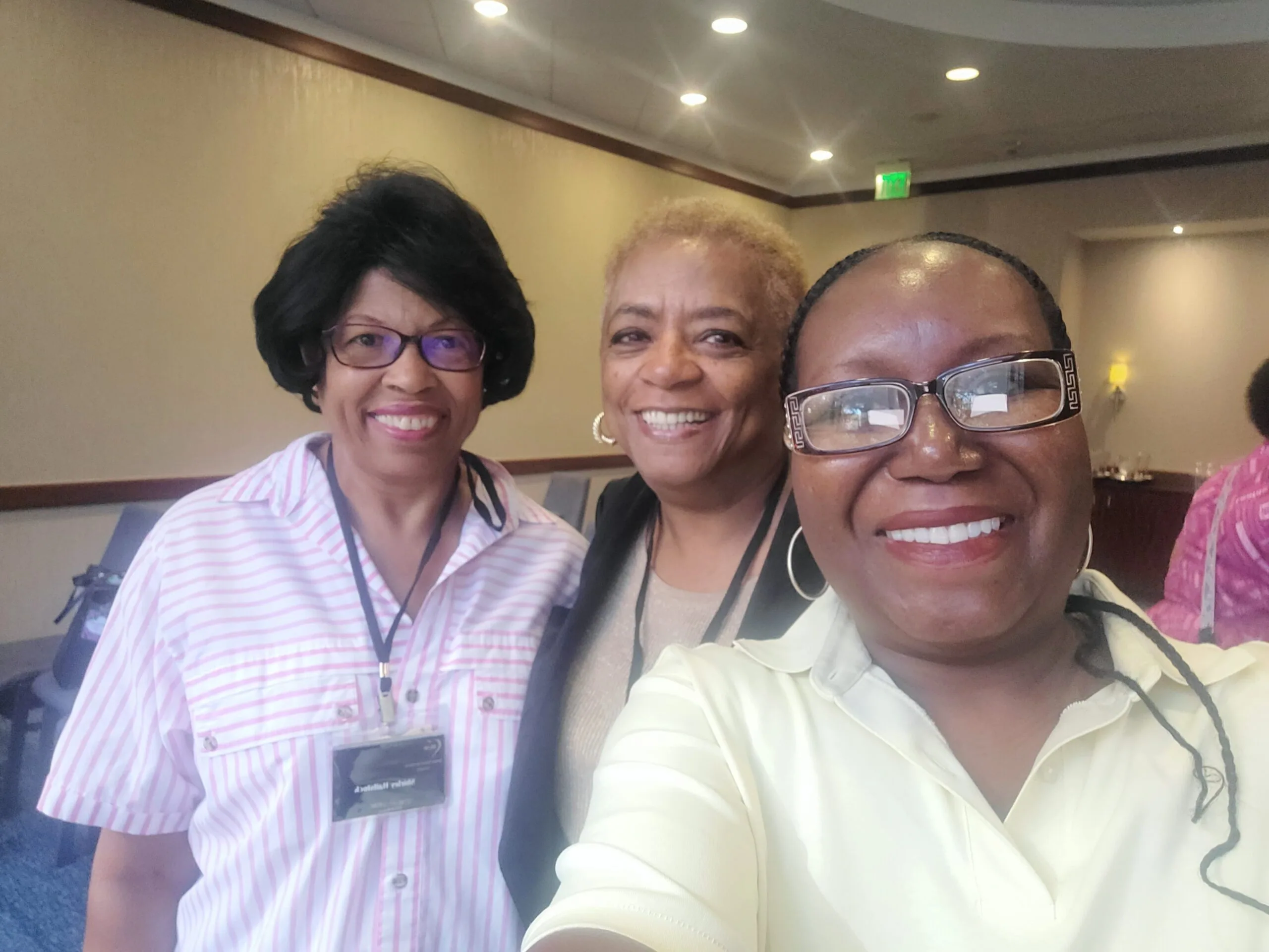 Three women smiling at a conference or event indoors, one wearing a badge and pink striped shirt, one in tan blazer, one in cream shirt with glasses